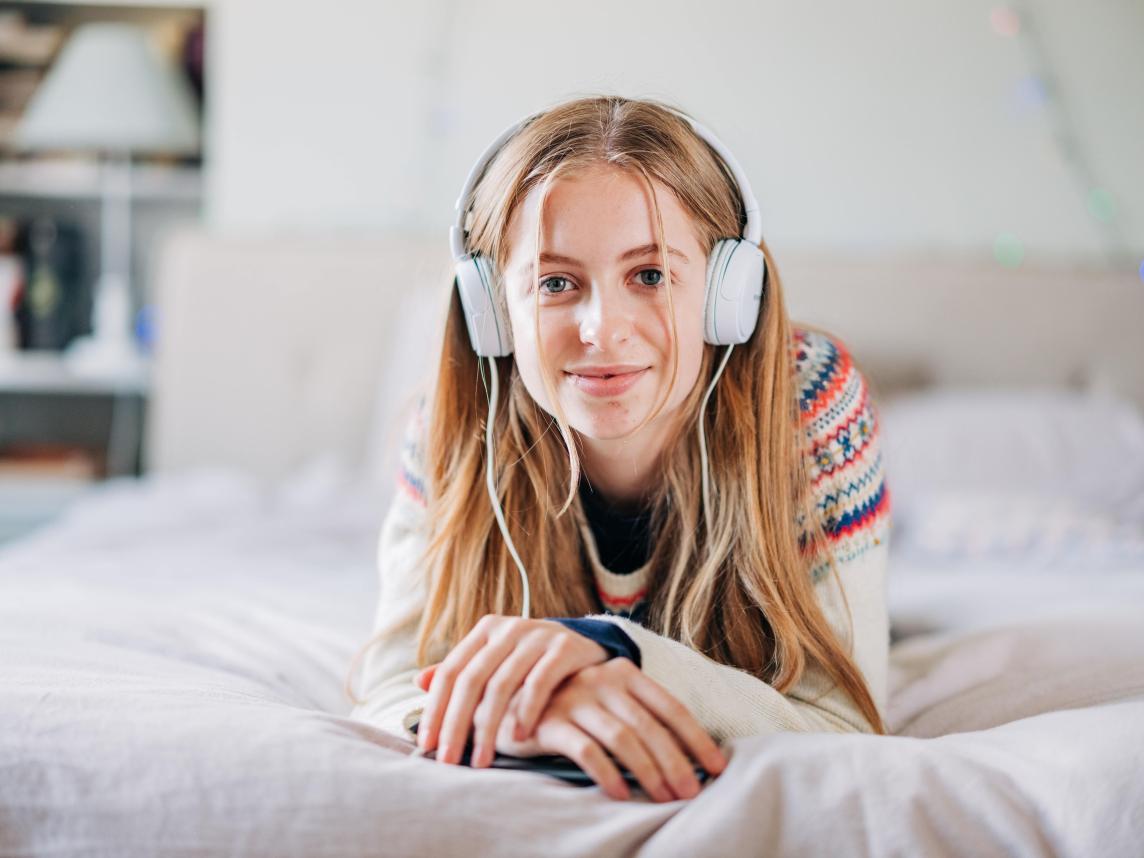 A happy teenager lies on her bed enjoying music looking directly at the camera