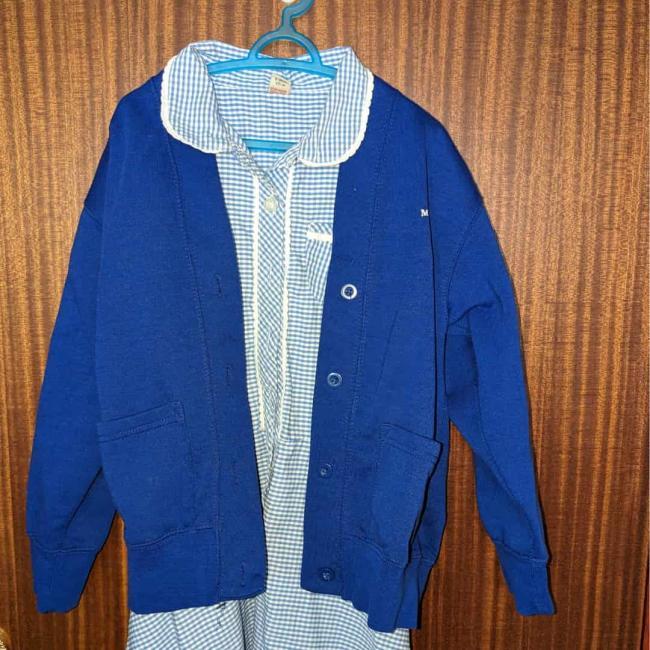 A blue checked girls school dress with a blue jumper on top, hanging on the back of a wooden door